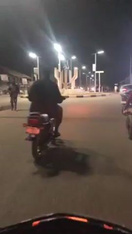 The video shows a first-person perspective of someone riding a motorcycle at night. The rider approaches a roundabout with several other motorcycles and pedestrians nearby. There are street lights illuminating the area and a tall monument in the center of the roundabout. The motorcycle rider passes through the roundabout, observing the other vehicles and people around. The dashboard of the motorcycle is visible, showing the speedometer.