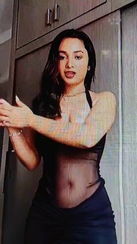 A young woman with long, dark hair is shown in a close-up shot. She is wearing a black, sheer dress with a halter neckline and a ruched skirt. She has a necklace and bracelets on. The background appears to be a closet or wardrobe with wooden doors and silver handles. The lighting is dim. The woman is looking at the camera and moving her arms. She touches her hair.