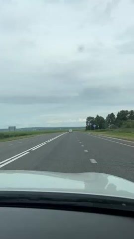 A vertical, first-person perspective video shows a white car driving down a two-lane asphalt road under a cloudy sky. The road has white dashed lines in the right lane and double solid white lines in the left lane. In the distance, two white cars are visible on the road. On the right side of the road, there are green trees and a small red structure. Two blue road signs are also visible on the right side of the road. The car is moving forward, and the scenery is passing by.