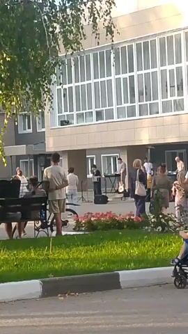 A group of people are gathered outdoors in front of a modern building with large windows. Some individuals are standing and observing a small band performing on a stage. The band consists of at least two musicians, one playing a keyboard and another holding a guitar. Other band members are visible behind them. Attendees are spaced out, with some standing and others seated on benches or in strollers. The environment appears to be a paved area with a lawn and some decorative plants in the foreground. The time of day seems to be late afternoon or early evening, given the lighting.