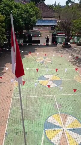 An aerial shot shows an outdoor courtyard decorated with red and white pennant flags. A flagpole with the Indonesian flag stands prominently in the foreground. The courtyard features a painted court with colorful circular designs. In the background, a person on a motorcycle enters through a gate, passing by some trees and buildings. The scene appears to be a schoolyard or a similar public space.