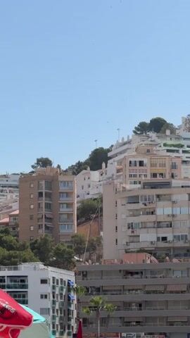 A vertical video captures a view of a densely packed hillside urban area under a clear blue sky. Buildings of varying heights and architectural styles, mostly apartments, are built on the slopes. The architecture appears predominantly modern, with many balconies visible. Some buildings are white, while others are cream or tan. Lush green trees are interspersed among the buildings, especially on the higher slopes. In the foreground, parts of what appear to be beach umbrellas and a person are visible, suggesting the viewpoint is from a beach or waterfront area. The overall impression is of a coastal city with a high density of residential buildings built into a hillside.