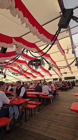 A video captures the interior of a large, tented venue, likely a beer hall or festival space. The ceiling is adorned with a festive decoration of alternating red and white ruffled fabric draped in a wavy pattern, interspersed with black streamers and some hanging wreaths. Speakers are visible attached to the ceiling structure. Long, communal tables with benches, all in a bright orange color, are set up in rows, with people seated at them. In the foreground, a woman and a man sit at one of these orange tables. The man is wearing traditional Bavarian attire, a dark vest over a white shirt. The woman is wearing a white shirt and a patterned skirt. They are engaged in conversation, with drinks on the table. Further back, numerous other people are seated at similar tables, some eating, some drinking, and some conversing, suggesting a lively atmosphere. The floor appears to be made of wooden planks. The overall environment suggests a cultural event or celebration, possibly Oktoberfest given the attire and decorations.