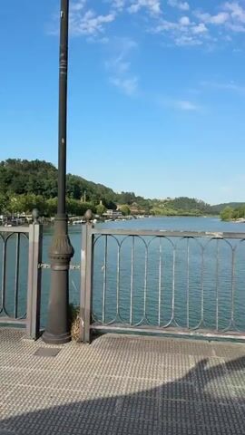 A woman wearing a black t-shirt, gray pants, and sandals walks across a bridge. She is carrying a water bottle in her left hand. The bridge has a metal railing with an ornate design. Beyond the railing is a body of water with several boats docked along the shore. In the background, there are green hills covered with trees and some buildings. The sky is blue with scattered white clouds. The sun is shining, casting shadows on the bridge.