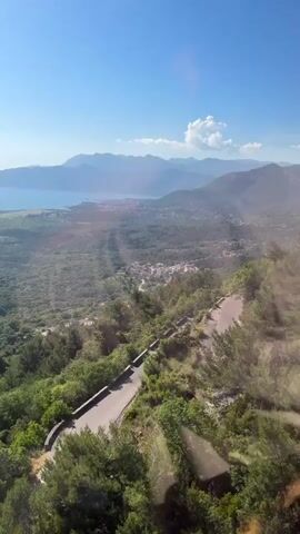 The video shows a high-angle view of a winding road cutting through a lush green landscape. In the distance, a body of water, possibly a bay or sea, stretches out under a clear blue sky. A town with numerous buildings is visible along the coastline. Mountains rise in the background, their peaks hazy in the distance. The sun is shining brightly, creating a slight glare.