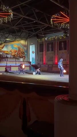 A live performance is taking place on a dirt arena floor, likely indoors due to the visible ceiling structure with lights and banners. In the background, a large mural depicts a chariot race. The arena is bordered by a low wall with decorative fabric draped over it. The performance appears to be a reenactment or show involving individuals in medieval-style costumes. In the foreground, on the left, a performer in a red tunic, black pants, and armor, holding a shield and a flaming sword, stands on the arena floor. To his right, another performer is kneeling, holding a shield. Further to the right, a third performer, dressed in a dark costume with a hood and mask, stands holding a sword. The lighting is dim with spotlights highlighting the performers. The audience area is not fully visible, but a glimpse of patterned fabric suggests seating or decorative elements in the foreground. The overall atmosphere suggests a staged event, possibly a jousting or gladiatorial show.