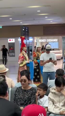 The video shows an indoor scene, possibly an airport or a public gathering space. In the background, a currency exchange counter is visible. Two young women in traditional Mongolian attire, featuring ornate robes and distinctive hats, stand near the counter. One woman wears a red and gold outfit, and the other wears an orange and black outfit. In the foreground, a group of people are seated, some appearing to be waiting. A man in a white t-shirt and jeans is looking at his phone. Several other individuals are also visible, including a woman in a teal shirt, a boy, and a person wearing a red baseball cap. The lighting suggests an indoor environment with overhead lights.