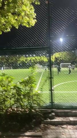 The video shows a nighttime scene of a soccer field, filmed from outside a chain-link fence with green foliage partially obscuring the view. The field is illuminated by bright overhead lights. Several people, appearing to be young men, are on the field, some walking and some actively playing soccer. There are multiple soccer goals visible. In the background, a multi-story building with illuminated windows is discernible behind the trees. Signs in what appear to be Chinese characters are attached to the chain-link fence. The camera moves slightly, shifting the view of the field.