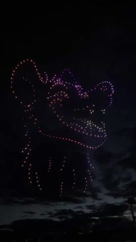A drone show in the night sky depicts the silhouette of a smiling lion. The drones are lit up in purple, orange, and white. Beneath the drone show are some clouds and a few lights in the distance.