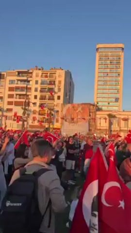 Here is a description of the video:

The video shows a crowd of people gathered in an open space, possibly a park or plaza, on a sunny day. They are waving Turkish flags, some of which bear the image of Mustafa Kemal Atatürk, the founder of modern Turkey. The atmosphere seems celebratory and patriotic, with people cheering and clapping. The scene is set against a backdrop of buildings, including a tall hotel and apartment complexes. The sky is clear and blue. The video captures a sense of national pride and unity among the crowd.