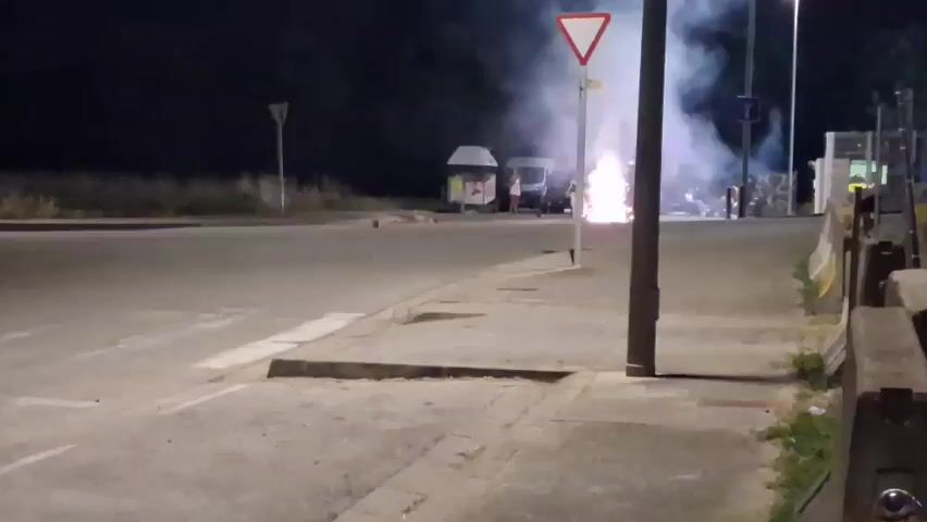 At night, on a paved area next to a road, several people are gathered around a bright, explosive firework. The firework is emitting sparks and smoke. There are a few vehicles in the background, including what appears to be a white van. There are also some small structures that might be kiosks or shelters. A yield sign is visible on a pole. The scene is lit by the firework and streetlights.