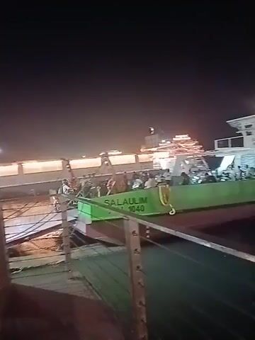At night, a ferry, named "SALAULIM 1040" with people on board, is docked at a pier. The ferry is green on the bottom and white on top. There is another structure on the pier decorated with lights. The water reflects the lights. The scene is dark. The camera is held by someone standing on the pier.
