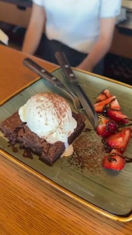 A person in a white shirt is seated at a wooden table, behind two dessert plates. The plate in the foreground on the left contains a chocolate brownie topped with a scoop of vanilla ice cream and dusted with cocoa powder. Sliced strawberries, drizzled with chocolate sauce, are on the right side of the plate. A knife and fork are positioned on top of the plate. The plate in the foreground on the right has a similar presentation of a brownie, ice cream, and strawberries.