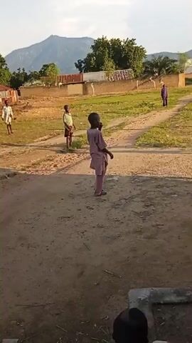 A group of children are playing soccer in a dirt field. The sun is shining and there are trees and buildings in the background. The children are wearing casual clothes and appear to be enjoying themselves. One child kicks the ball, and another child runs to try and intercept it. The game is informal, with no referee or goals.