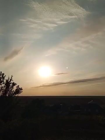 The video captures a rural landscape at sunset. The sun, a bright orb, is visible in the upper left quadrant of the frame, casting a warm glow across a pale blue sky streaked with wispy clouds. Below the sky, a dark horizon line separates the sky from the land. The land itself is a muted silhouette, featuring a few houses with visible roofs and some trees. The foreground is dominated by dark foliage, suggesting plants or bushes. The camera slowly pans to the right, revealing more of the landscape and the sky. The overall impression is one of tranquility and the end of a day in a peaceful, natural setting.