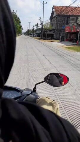The video shows a first-person perspective of someone riding a motorcycle down a paved road. The road is marked with white lines. On the right side of the road, there are buildings, some of which appear to be shops or businesses. One building has a red sign. There are power lines overhead. A white car is parked on the right side of the road. A few trees are visible along the side of the road. The sky is blue with some clouds. The rider's hand and part of the motorcycle's handle are visible in the foreground. The video appears to be taken during the daytime.