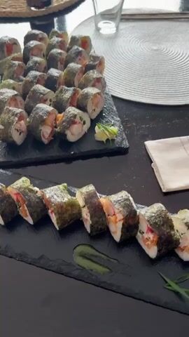 The video shows two black rectangular serving platters filled with sushi rolls. The upper platter, placed further back and slightly higher, holds approximately 20 sushi rolls arranged in rows. These rolls are wrapped in dark green seaweed and appear to contain rice, pinkish-orange ingredients, and possibly white seafood or vegetables. To the right of this platter, a small mound of finely shredded pickled ginger is visible. The lower platter, closer to the camera and at a lower level, contains about 7-8 sushi rolls, similarly wrapped in seaweed and showing similar fillings. A swirled green sauce, possibly wasabi or an herb-based sauce, is drizzled on the platter in front of these rolls. The serving platters rest on a dark wooden surface. In the background, a portion of a white circular placemat and a clear glass can be seen. The lighting is natural and appears to be from above.