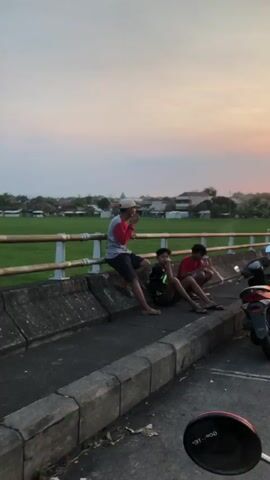 Three young males are sitting on a concrete barrier next to a metal railing. The person on the left is wearing a red and grey long-sleeved shirt, a white hat, and dark shorts. They are holding something to their face. The person in the middle is wearing a black t-shirt with green and yellow accents and dark shorts. They have their hand resting on their chin. The person on the right is wearing a red t-shirt and dark shorts. They are crouching and holding something to their face. The ground is paved, and there is a motorcycle partially visible in the lower right corner. Behind the barrier is a green field, possibly rice paddies, with some buildings and trees in the distance. The sky is overcast with hints of color, suggesting it is either sunrise or sunset.