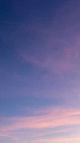 The camera looks up at a colorful sky. The sky is a blend of purple, blue, and pink, with the pink hues concentrated near the bottom of the frame. A faint contrail is visible in the upper-left. A mountain is barely visible at the bottom of the frame.