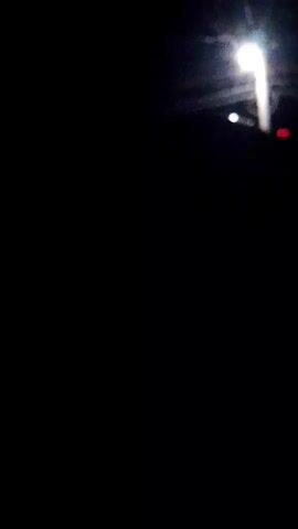 The video is a very dark, blurry recording of a street lamp at night. The lamp is white and bright, and it appears to be attached to a pole. There are some power lines visible in the background, as well as some faint lights that could be other street lamps or lights from buildings. The camera is shaking slightly, making the image even more unclear. It is difficult to discern any specific details or actions due to the poor quality of the recording.