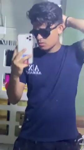 A young man in a dark blue Armani Exchange t-shirt and dark pants takes a selfie in front of a mirror, wearing dark sunglasses. He adjusts his hair and points to himself in the reflection. The background includes a cabinet with a white frame and various objects. The lighting is somewhat dim.