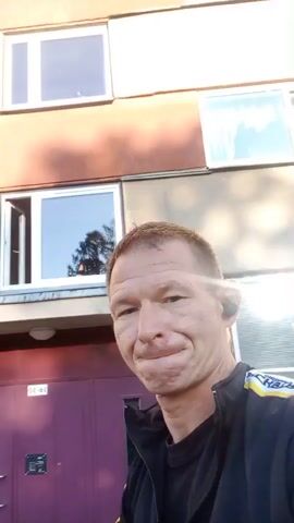 A man with short brown hair and a light complexion is standing outside a building with a reddish-brown facade and several windows. He is wearing a black jacket with yellow and blue accents. The man is looking at the camera with a neutral expression. The video is shaky, suggesting it was filmed by the man himself. The lighting indicates it is daytime, possibly late afternoon given the angle of the sun creating shadows. The building appears to be an apartment complex or similar multi-story structure.