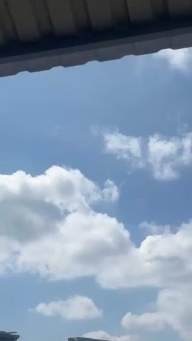 The video shows a blue sky with scattered white clouds. The top of the frame is partially obscured by a dark, corrugated roof or overhang. A small, dark object, possibly a bird or insect, is visible in the sky in the upper left quadrant of the frame. The clouds are moving and changing shape as they drift across the sky. In the lower portion of the frame, the tops of some buildings are visible. The scene appears to be outdoors on a clear, possibly sunny day.