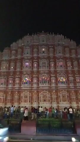 At night, the Hawa Mahal in Jaipur, India, stands grandly, illuminated in warm light, showcasing its many windows. Pedestrians stroll along the sidewalk in front of the building, some pausing to take photographs. Cars pass by in the foreground, creating movement and a dynamic atmosphere. The sky above is dark, emphasizing the architectural details of the building and the street-level activity.