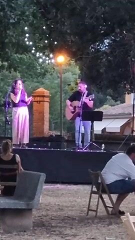A man plays guitar and sings on a stage outdoors. A woman stands next to him and sings into a microphone. A single lamp illuminates the stage from behind them. Numerous small lights hang in the trees above. Several people are seated in chairs and on a bench in front of the stage, watching the performance. The ground appears to be dirt and grass.