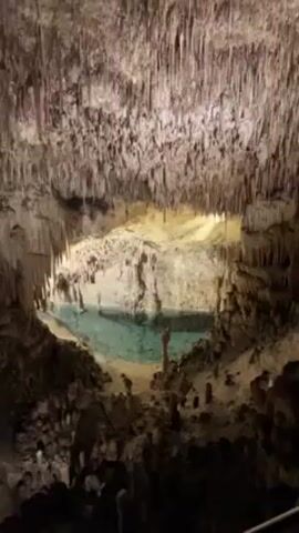 The video shows the interior of a cave featuring a pool of clear, turquoise-colored water. The cave is illuminated, highlighting stalactites hanging from the ceiling and stalagmites rising from the cave floor. The walls are textured and rocky, creating a rugged and cavernous environment. The camera pans slightly, offering a wider view of the cave's interior, revealing more details of the rock formations and the unique geological features of the cave.