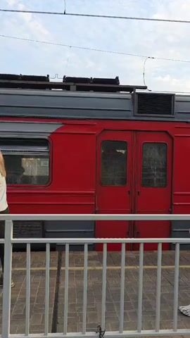 A young woman with brown hair tied in a ponytail, wearing a dark hoodie, grey sweatpants, and white Crocs, walks past a red train car holding a white cup and a white tote bag. Behind her, the train doors open and close. A second woman with short blonde hair, wearing a teal and white tracksuit, approaches a suitcase. She is standing on a train platform with a white railing in the foreground. The sky is cloudy.