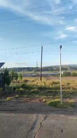 The video shows a street with power lines overhead, a building, and a boy walking on a path. The boy is wearing a light gray shirt and dark shorts. He walks from the right side of the screen to the left, then out of frame. The street is asphalt, and there are grassy areas on either side. In the background, there are hills and some buildings. The sky is blue with some clouds.
