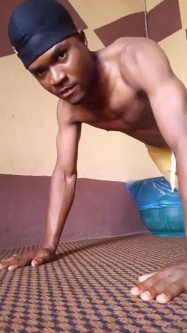 A shirtless Black male in black head covering is doing pushups. He is wearing yellow athletic shorts with white stripes. His hands are on the floor on a patterned brown carpet. A teal laundry basket with a blue trash bag is in the background, against a brown and tan paneled wall. The man does a series of pushups.