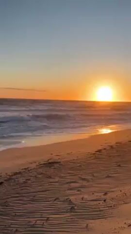 The video shows a beach at sunset. The sun is setting over the ocean horizon, casting an orange glow on the sky and water. Gentle waves are coming ashore, reflecting the light. The sand is a mixture of golden and reddish hues, marked with ripples and some footprints. The sky transitions from a light blue overhead to orange near the horizon. There are no people visible in the shot.