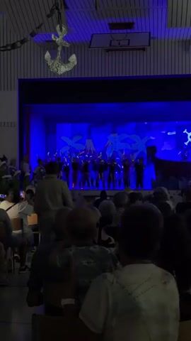 A group of children in costumes are performing on a stage in front of a blue backdrop. Some of the children are dressed as pirates, as indicated by the pirate flag visible on the backdrop. The audience, composed of adults and children, is seated in chairs facing the stage. The room appears to be a gymnasium or auditorium, with a basketball hoop visible on the ceiling. Decorative anchors hang from the ceiling, suggesting a nautical theme. The lighting is dim, with blue stage lights illuminating the performance.