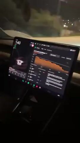 A video shot from inside a car at night shows the digital dashboard speedometer rapidly increasing from 141 to 164. To the right of the speedometer is a graph displayed on the dashboard showing the car's energy usage. There is also music playing on the dashboard screen. Outside of the car, there are trees and street lights.
