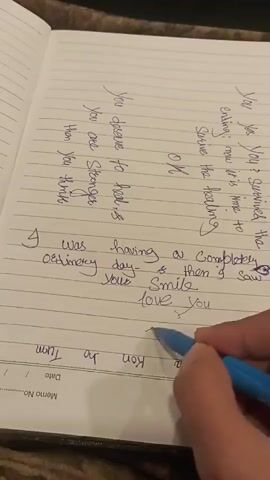 A close-up shot shows a person's right hand holding a blue pen, writing on a lined notebook. The notebook is open to a page with handwritten text in purple ink. The writing is in cursive and some parts are upside down and backwards. The text appears to be a poem or a personal reflection. The background is blurry and appears to be a table with a textured surface. The lighting is natural and coming from the top left.
