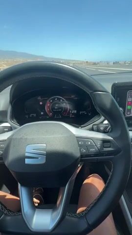 A first-person perspective video shows the interior of a car driving on a sunny day. The driver's hands are visible on the steering wheel, and the car's dashboard displays a digital speedometer at 125 and navigation information on the central screen. The road ahead is a highway, with the ocean visible to the right. The sky is clear blue.