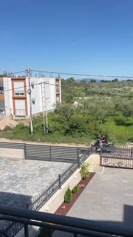 A person rides a scooter down a paved path, past a white building with balconies. A black metal fence runs along the path. In the background, there are trees and more buildings. The sky is blue.