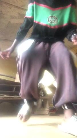 A young boy dances in front of a camera. He is wearing a black long-sleeved shirt with green and red stripes across the chest, and purple pants. He is in a room with a concrete ceiling, wooden furniture, and a purple plastic chair. The boy is in motion, and his feet are blurred.