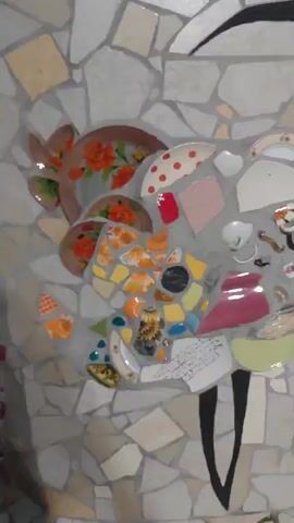 This is a vertical video of a mosaic artwork depicting a bird. The mosaic is made of various broken ceramic pieces, including plates, cups, and tiles, arranged to form the shape of a crane or stork with its wings outstretched. The bird's body is filled with a collage of these ceramic fragments in different colors and patterns. Some visible fragments include pieces with floral designs, red polka dots, and solid colors like yellow, blue, and red. There are also some smaller objects embedded in the mosaic, such as what appear to be small figurines or charms. The background of the mosaic is composed of larger, irregular-shaped light-colored tiles. The bird's legs are depicted as two black, narrow shapes extending downwards. To the right of the bird's head, there is a small rectangular mosaic detail with green and black tiles, resembling a windowpane. The lighting appears to be overhead, casting a slight shadow on the artwork.