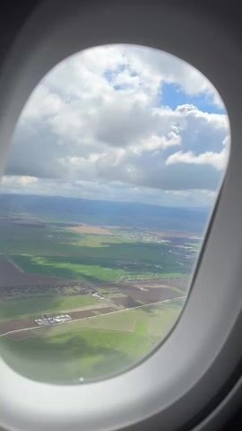 The view from a plane window shows a patchwork of green and brown fields below, with buildings clustered in small settlements. The sky is partly cloudy, with patches of blue visible between the puffy white clouds. A hazy outline of mountains can be seen in the distance. The plane window frame is visible around the edges of the shot.
