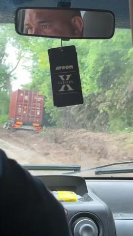 The video, shot from inside a vehicle, shows a challenging road condition in what appears to be a rural or semi-rural area, consistent with the given Dalgopol, Bulgaria context. The primary focus is a large red semi-trailer truck with a cargo container, which is stuck in a substantial amount of mud and debris on the side of the road.

The camera's perspective is from the front passenger seat of a car, looking out through the muddy windshield. The car's dashboard is visible at the bottom of the frame. A rearview mirror hangs centrally, reflecting a portion of the driver's face. Hanging from the rearview mirror is a black rectangular air freshener with the brand "areon" and "X VERSION" printed on it in white text.

The road itself is severely affected by mud, suggesting heavy rainfall, flooding, or a mudslide. Large piles of reddish-brown mud and debris are visible on both sides of the road and partially covering the asphalt or unpaved surface. The truck's wheels are deeply embedded in this mud, and its trailer is tilted towards the right, indicating it has slid off the main road or is struggling to gain traction. The back of the red container has some text, partially visible as "TIRU 41GP-GRP" and "45G1".

Lush green trees and foliage line the road, and a power line tower can be briefly seen in the background on the left. The sky is overcast, suggesting daytime with poor weather conditions. The overall scene depicts a difficult and potentially hazardous travel situation due to the adverse environmental conditions.