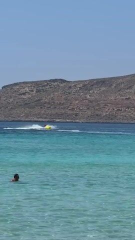 A jet ski is being ridden by two people in the blue sea. A yellow buoy is to the left of the jet ski. A rocky hill is in the background. The sky is clear and blue.