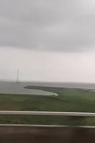 The video shows a landscape view, likely taken from a moving vehicle, featuring a large expanse of water under a cloudy sky. Two electrical transmission towers are visible in the distance, standing in the water. In the foreground, there is a stretch of green land. The camera is positioned behind a barrier, probably a roadside guardrail, visible at the bottom of the frame. Occasional reflections appear on the window through which the scene is being viewed, and there is some minor distortion due to the window’s shape or imperfections.