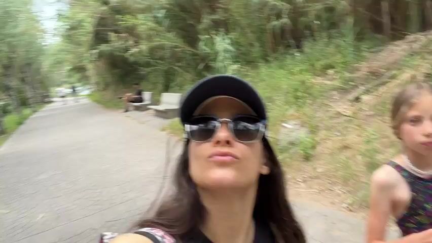 A woman is recording a video while walking down a park path. The path is paved with a circular pattern and lined with trees on one side and dense foliage on the other. A man is sitting on a bench further down the path. Two people are jogging in the distance. The camera is held close to the woman's face, showing her wearing sunglasses and a baseball cap. A river runs alongside the path. The sky appears overcast.