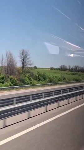 This is a short video clip, seemingly taken from a vehicle, offering a view of a highway with farmland and a clear blue sky. 

The main subjects are the highway itself, with its asphalt surface and white lane markings, and the protective barriers separating the lanes. Beyond the barriers, lush green fields stretch towards a line of trees and a few scattered bare trees. The sky is a vibrant, cloudless blue, suggesting a sunny day. Reflections are visible on the window through which the video is filmed, indicating the perspective of someone traveling.

The activity is travel along a highway. The video captures the passage of at least two vehicles: a white car and a dark-colored car. The time of day appears to be daytime, given the bright sunlight and clear sky. The weather seems pleasant and fair. While no specific landmarks are immediately identifiable, the presence of agricultural fields and the highway suggests a rural or semi-rural setting. The overall mood is one of motion and a journey through a serene landscape. There is no visible text in the clip. The provided location context "Malko Tranovo, Bulgaria" helps to place the scene geographically.