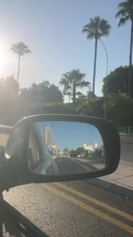 The video is a first-person perspective from inside a vehicle, looking at the side-view mirror. The mirror reflects a street scene with buildings and palm trees under a bright, sunny sky. The vehicle appears to be moving, as indicated by the slightly blurred foreground and the shifting perspective in the mirror. The road in the reflection has yellow lane markings. The overall environment appears to be a warm, possibly tropical or Mediterranean location. There are no people visible in the frame, only the car interior, the side-view mirror, and the reflected scenery.