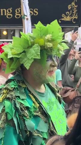 The video shows a person dressed as the Green Man standing in a crowd of people, some of whom are taking pictures of him. He has green face paint and a beard, and wears a leafy crown and a garment made of green fabric and leaves. Behind him, there is a woman with a colorful flower crown. There is also a shop sign that says "ing glass" visible in the background.