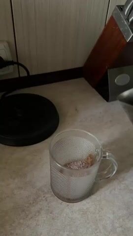In a close-up shot, hot water is being poured from a metal kettle into a clear glass mug filled with a brown powder, likely coffee or hot chocolate. The mug is on a speckled countertop. In the background, a black electric appliance, possibly a kettle base, is visible to the left, and a knife block with wooden-handled knives stands to the right. The scene is set in what appears to be a kitchen, with tiled walls in the background. The water stream creates froth as it mixes with the powder.